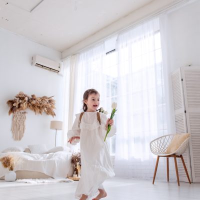 Happy little girl in flowing white dress runs barefoot in bright, sunlit room. Airy space, interior decorated in minimalist style, enhances feeling of freedom, joy. Happiness childhood. Boho style